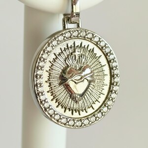 May include: A circular silver-toned pendant featuring a central embossed Sacred Heart design with radiating sunburst lines. The outer edge of the pendant is framed by a border of small, clear, sparkling stones set into the metallic surface.