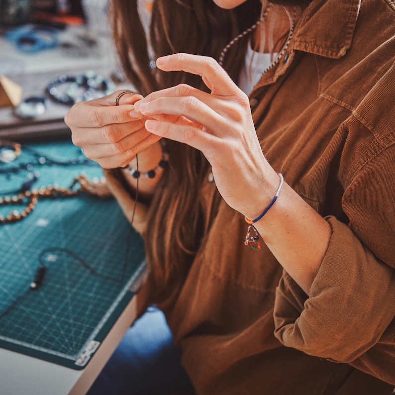 May include: A beaded bracelet featuring a thin blue cord, a white segment, and a small multi-colored charm. The bracelet is worn on a wrist, with the hands actively working on a thin dark string, while a brown corduroy shirt is visible.