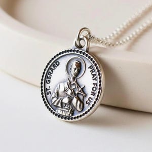 May include: A silver-colored pendant necklace featuring a detailed depiction of St. Gerard. The round pendant has the words "ST. GERARD" and "PRAY FOR US" around the edge. The saint is holding a cross. The necklace chain is a delicate beaded style.