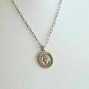 May include: A silver-toned necklace featuring an oval pendant with a raised Sacred Heart emblem surrounded by a sunburst pattern and a beaded border. The pendant hangs from a delicate chain composed of elongated, rectangular-shaped metal links.