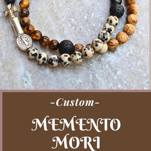 May include: A collection of three beaded rosary bracelets featuring various natural stone beads, including black lava, dalmatian jasper, and tiger eye. Each bracelet incorporates a metal crucifix charm and a small circular religious medal.
