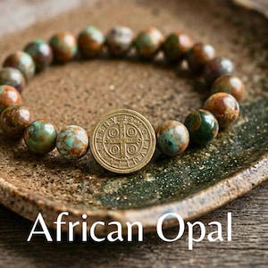 Puede incluir: Una pulsera de cuentas con piedras redondas de ópalo africano multicolor en tonos verdes, marrones y turquesas. La pulsera incluye un dije central circular de metal color latón grabado con una cruz y las letras V R S N S M V S M Q L I V B.
