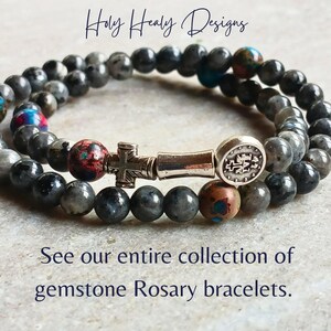 May include: A double-wrap gemstone rosary bracelet featuring dark grey beads, interspersed with multi-colored speckled beads, a silver-toned cross charm, a cylindrical spacer, and a round medal. Text reads: Holy Healy Designs. See our entire collection of gemstone Rosary bracelets.