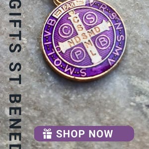 May include: A collection of gold-tone Saint Benedict pendant necklaces featuring circular medals with colorful enamel inlays in shades of red, purple, and blue, suspended from delicate gold-tone link chains on a textured stone surface.