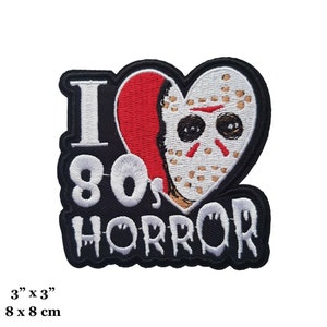 Puede incluir: Parche bordado en blanco y negro con un corazón rojo. El corazón presenta una máscara de hockey blanca con un corte sangriento en la cara. El texto "I 80's HORROR" está bordado en blanco sobre un fondo negro.