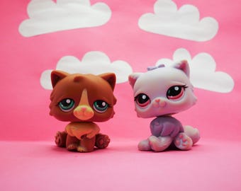 Puede incluir: Dos pequeños gatos de juguete de plástico. Un gato es marrón con marcas blancas y el otro es morado con marcas blancas. Ambos gatos tienen ojos grandes y están sentados sobre una superficie rosa.