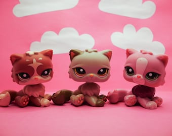 May include: Three small plush toy cats with different colored fur. The cat on the left is brown with white markings, the middle cat is gray with white markings, and the cat on the right is pink with white markings.