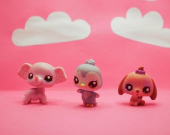 May include: Three small plastic figurines of animals. A pink elephant, a blue penguin, and a brown dog. The figurines have large eyes and are set against a pink background.