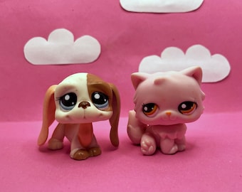 Puede incluir: Dos pequeñas figuras de animales de plástico: un perro y un gato. El perro es blanco y marrón con grandes ojos azules. El gato es rosa con ojos naranjas. Ambos están sobre un fondo rosa con nubes de papel blancas.