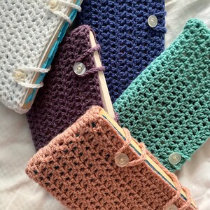 May include: Five crocheted book covers in different colors, including white, purple, blue, green, and pink. Each cover has a button closure.