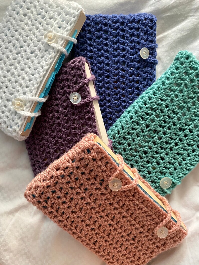 May include: Five crocheted book covers in different colors, including white, purple, blue, green, and pink. Each cover has a button closure.