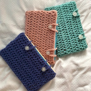 May include: Three crocheted book covers in different colors: blue, pink, and green. Each cover has a button closure and a simple, open-weave design.