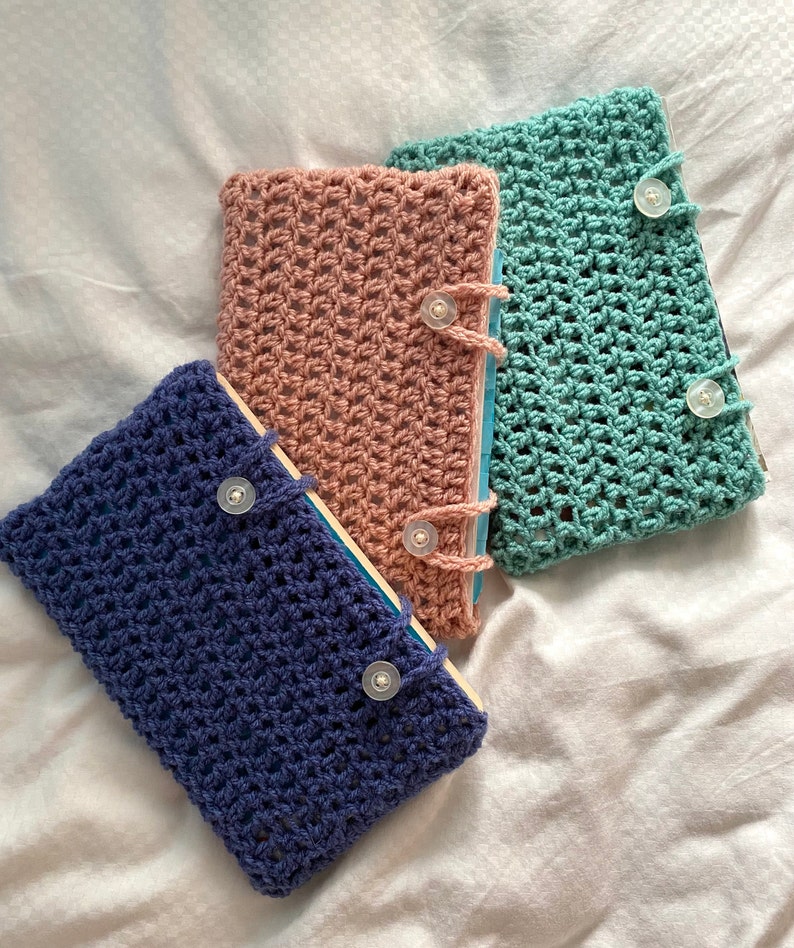 May include: Three crocheted book covers in different colors: blue, pink, and green. Each cover has a button closure and a simple, open-weave design.