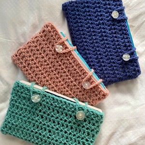 May include: Three crocheted book covers in different colors: teal, pink, and blue. Each cover has a button closure and a woven pattern.