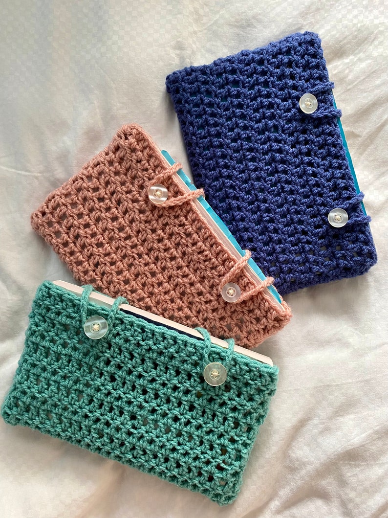 May include: Three crocheted book covers in different colors: teal, pink, and blue. Each cover has a button closure and a woven pattern.
