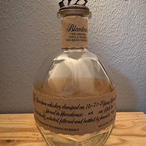 May include: A clear glass Blanton's bourbon whiskey bottle with a faceted, rounded body and a tan paper label. The bottle features a metallic stopper shaped like a horse and jockey. The label text includes "Blanton's The Original Single Barrel Bourbon Whiskey" and "Bourbon whiskey dumped on 10-21-21".