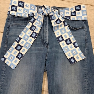 May include: Blue denim jeans with a decorative belt. The belt is white with a repeating pattern of blue squares and the letters "NC". The belt is tied in a bow in the front.
