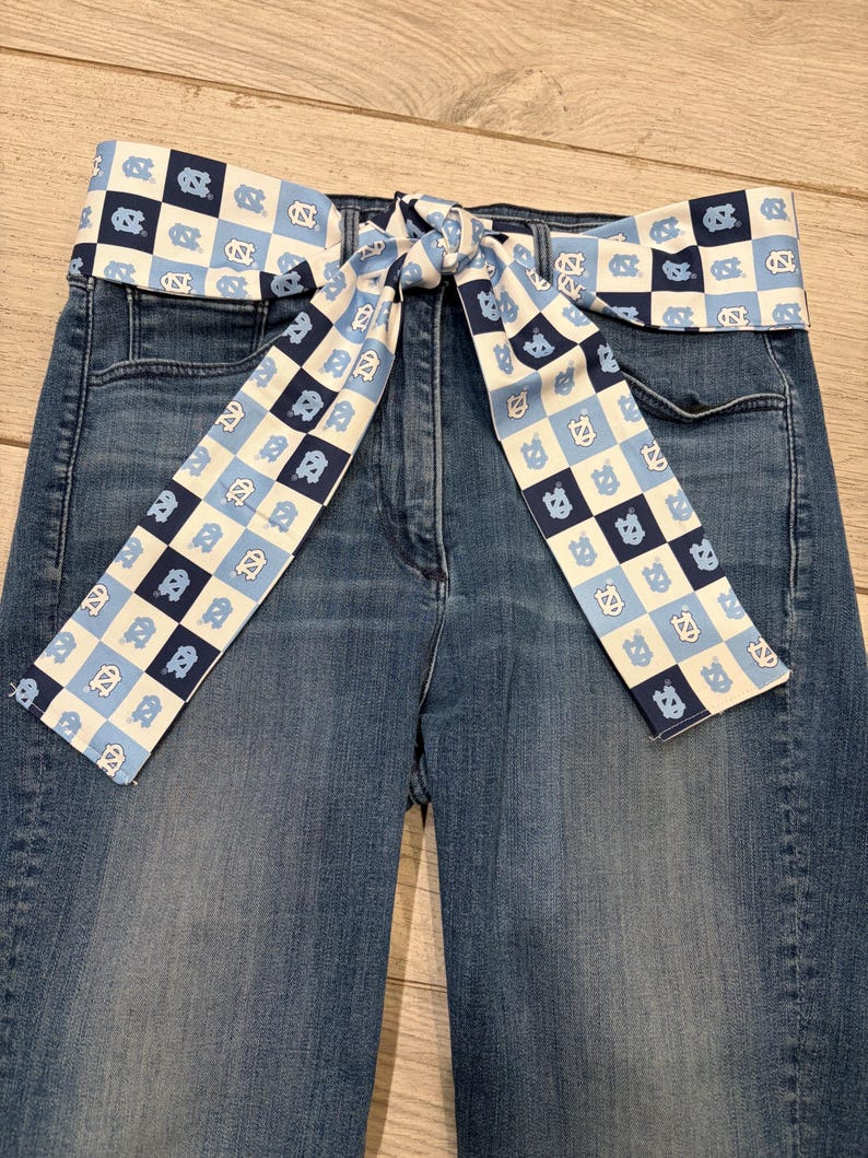 May include: Blue denim jeans with a decorative belt. The belt is white with a repeating pattern of blue squares and the letters "NC". The belt is tied in a bow in the front.