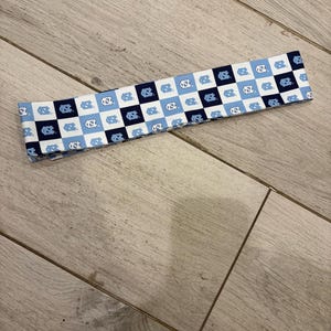 May include: A fabric headband featuring a checkered pattern of white, light blue, and navy blue squares. Each square contains a stylized logo. The headband is laying on a light wood surface.