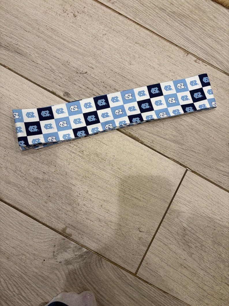 May include: A fabric headband featuring a checkered pattern of white, light blue, and navy blue squares. Each square contains a stylized logo. The headband is laying on a light wood surface.