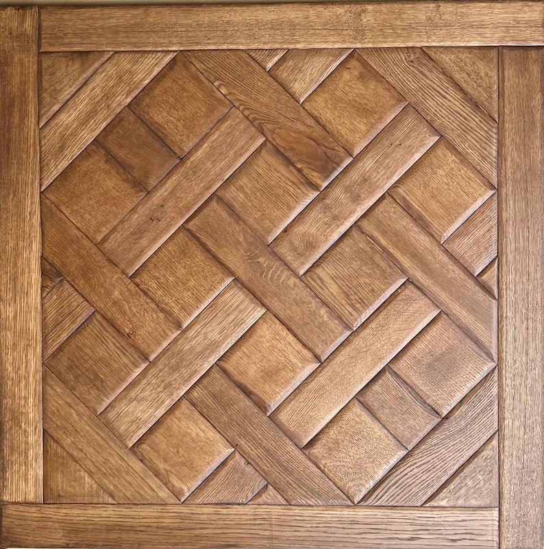 May include: A square wooden panel with a parquet pattern. The panel features a repeating herringbone design in various shades of brown. The outer edge is framed with a darker wood border, creating a defined edge.