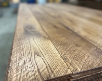 May include: Close-up of stacked, unfinished hardwood flooring planks. The planks are a warm, natural wood tone with visible grain and texture. The wood has a light brown color with darker brown streaks.