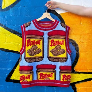Op de afbeelding: Een sleeveless gebreide vest met een blauwe achtergrond en een patroon van pindakaaspotten. De potten zijn rood, geel en bruin met het woord "Peanut" erop gedrukt.