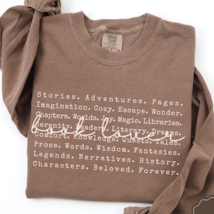 May include: Espresso-colored long-sleeve shirt with the words "Book Lover" in a cursive font. The shirt features a list of words related to reading, such as "Stories," "Adventures," and "Imagination." The shirt has a crew neck and a Comfort Colors tag.