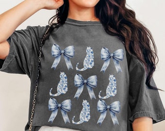May include: A dark gray t-shirt with a pattern of blue bows and white cats. The cats are outlined in blue and have a floral pattern inside.