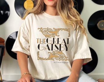 May include: Ivory-colored t-shirt featuring the text "THE GREAT GATSBY" in black and gold, with decorative flourishes. The author's name, F. Scott Fitzgerald, is printed below. The shirt is a relaxed fit.