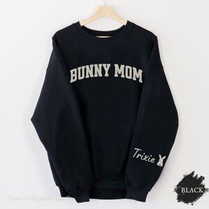Peut inclure: Sweat-shirt noir avec le texte "BUNNY MOM" en blanc sur le devant et le texte "Trixie" avec un graphique de lapin en blanc sur la manche gauche.