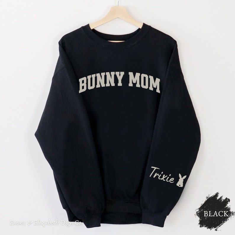 Peut inclure: Sweat-shirt noir avec le texte "BUNNY MOM" en blanc sur le devant et le texte "Trixie" avec un graphique de lapin en blanc sur la manche gauche.