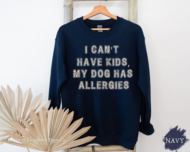 May include: Navy blue sweatshirt with the text "I CAN'T HAVE KIDS, MY DOG HAS ALLERGIES" in white. The sweatshirt is hanging on a wooden hanger against a white background. The brand name "Emma & Elizabeth Dsgn Co." is at the bottom.