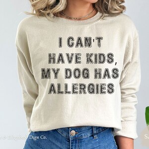 May include: A sand-coloured sweatshirt featuring the black text: "I CAN'T HAVE KIDS, MY DOG HAS ALLERGIES". The crew neck sweatshirt is a neutral colour. The brand name "Emma & Elizabeth Dsgn Co." is at the bottom left.