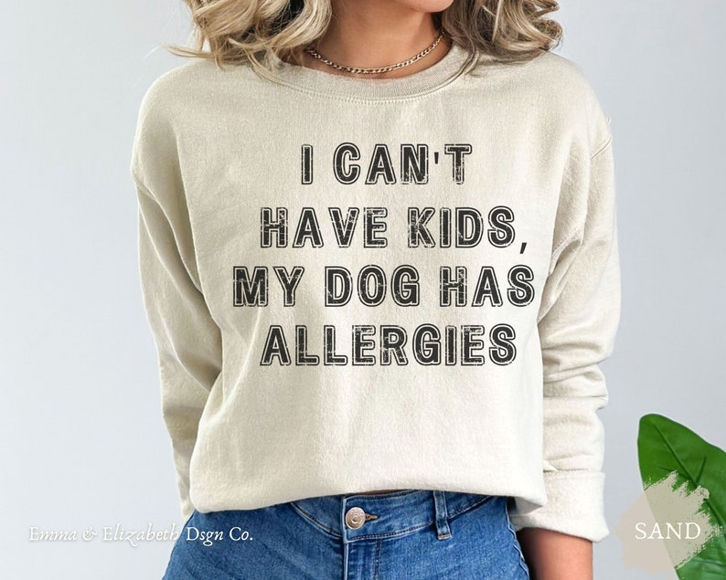 May include: A sand-coloured sweatshirt featuring the black text: "I CAN'T HAVE KIDS, MY DOG HAS ALLERGIES". The crew neck sweatshirt is a neutral colour. The brand name "Emma & Elizabeth Dsgn Co." is at the bottom left.