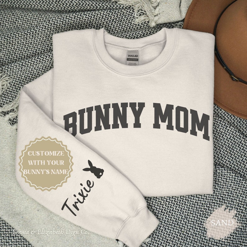 Peut inclure: Un sweat-shirt beige clair avec le texte "BUNNY MOM" en noir sur le devant. La manche gauche a une silhouette de lapin noir et le texte "Trixie" en noir. Une &eacute;tiquette blanche avec le texte "CUSTOMIZE WITH YOUR BUNNY'S NAME" est sur la manche gauche. Le texte "Emma & Elizabeth Design Co." est en bas &agrave; gauche du sweat-shirt.