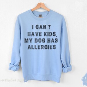 May include: Pale blue sweatshirt featuring the text "I CAN'T HAVE KIDS, MY DOG HAS ALLERGIES" in black. The garment is displayed on a wooden hanger. The words "LIGHT BLUE" are in the bottom right corner.