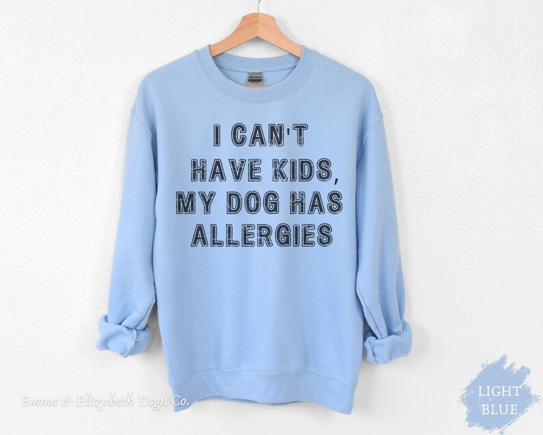 May include: Pale blue sweatshirt featuring the text "I CAN'T HAVE KIDS, MY DOG HAS ALLERGIES" in black. The garment is displayed on a wooden hanger. The words "LIGHT BLUE" are in the bottom right corner.