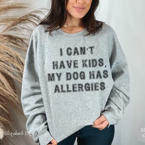 May include: A grey crew neck sweatshirt with the slogan "I CAN'T HAVE KIDS, MY DOG HAS ALLERGIES" printed in black. The sweatshirt is a casual style with long sleeves. The brand name "Emma & Elizabeth Dsgn Co." is printed at the bottom left.