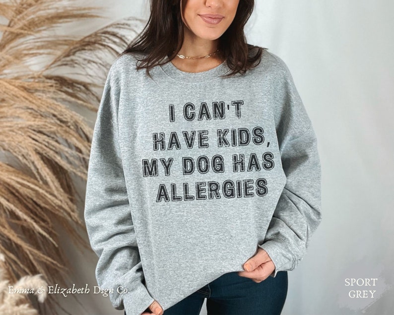 May include: A grey crew neck sweatshirt with the slogan "I CAN'T HAVE KIDS, MY DOG HAS ALLERGIES" printed in black. The sweatshirt is a casual style with long sleeves. The brand name "Emma & Elizabeth Dsgn Co." is printed at the bottom left.