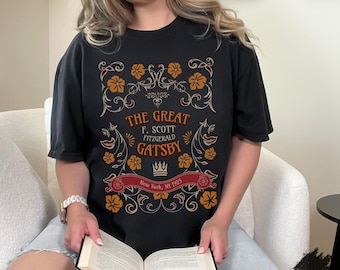 May include: A black t-shirt with a vintage-style graphic design featuring the text "The Great F. Scott Fitzgerald Gatsby" and "New York, NY 1925". The graphic includes a crown, floral elements, and a scroll.
