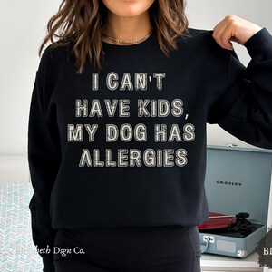 May include: Black sweatshirt featuring the text "I CAN'T HAVE KIDS, MY DOG HAS ALLERGIES" in a distressed, off-white font. The sweatshirt has a crew neck and long sleeves. The word "BLACK" is in the lower right corner.