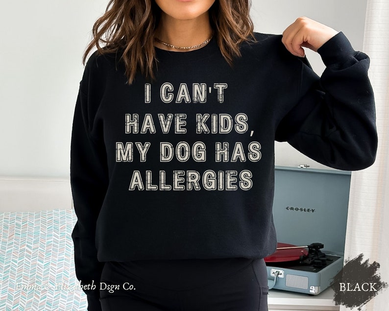May include: Black sweatshirt featuring the text "I CAN'T HAVE KIDS, MY DOG HAS ALLERGIES" in a distressed, off-white font. The sweatshirt has a crew neck and long sleeves. The word "BLACK" is in the lower right corner.