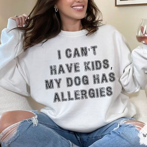 May include: Cream-coloured sweatshirt featuring the text "I CAN'T HAVE KIDS, MY DOG HAS ALLERGIES" in black. The sweatshirt has a crew neck and long sleeves. The word "WHITE" is in the bottom right corner.