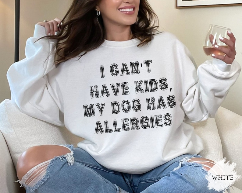 May include: Cream-coloured sweatshirt featuring the text "I CAN'T HAVE KIDS, MY DOG HAS ALLERGIES" in black. The sweatshirt has a crew neck and long sleeves. The word "WHITE" is in the bottom right corner.