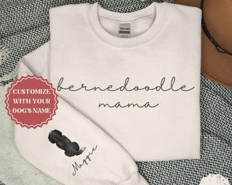 May include: A white sweatshirt with the text "bernedoodle mama" in black script. The sweatshirt has a black and white doodle of a dog on the sleeve with the name "Maggie" written underneath.