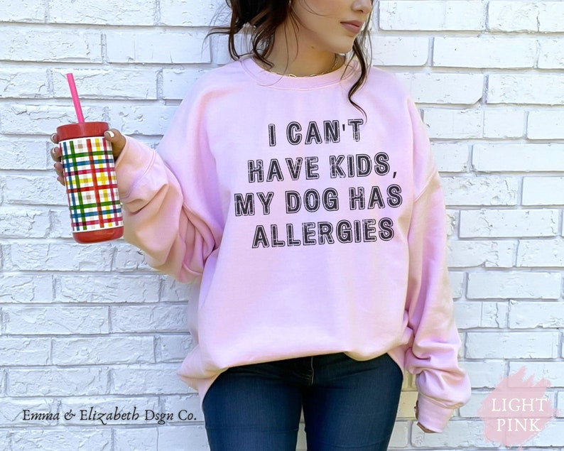 May include: Pale pink sweatshirt featuring the text "I CAN'T HAVE KIDS, MY DOG HAS ALLERGIES" in black. A person holds a tumbler with a red lid and a colourful plaid design. The words "Emma & Elizabeth Dsgn Co." are at the bottom.