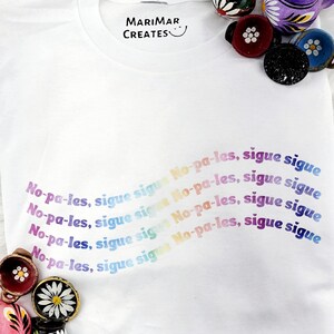 May include: White t-shirt with the text "No-pa-les, sigue sigue" repeated in a rainbow gradient. The text is in a playful, hand-drawn style. The shirt is a blank canvas for a fun and colorful design.