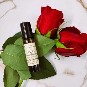 May include: A small, brown glass perfume bottle with a black cap, labeled "SAVIA SUGAR CANE PERFUME", rests on green leaves next to two vibrant red roses. The bottle contains 10 ml of fragrance oil. The background is a white marble surface.