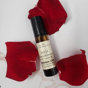 May include: A roll-on perfume bottle with a black cap and a label that reads "SAVIA LUX Sugar Cane Perfume Forbidden Love." The bottle is surrounded by three bright red rose petals on a white and gray marble surface.